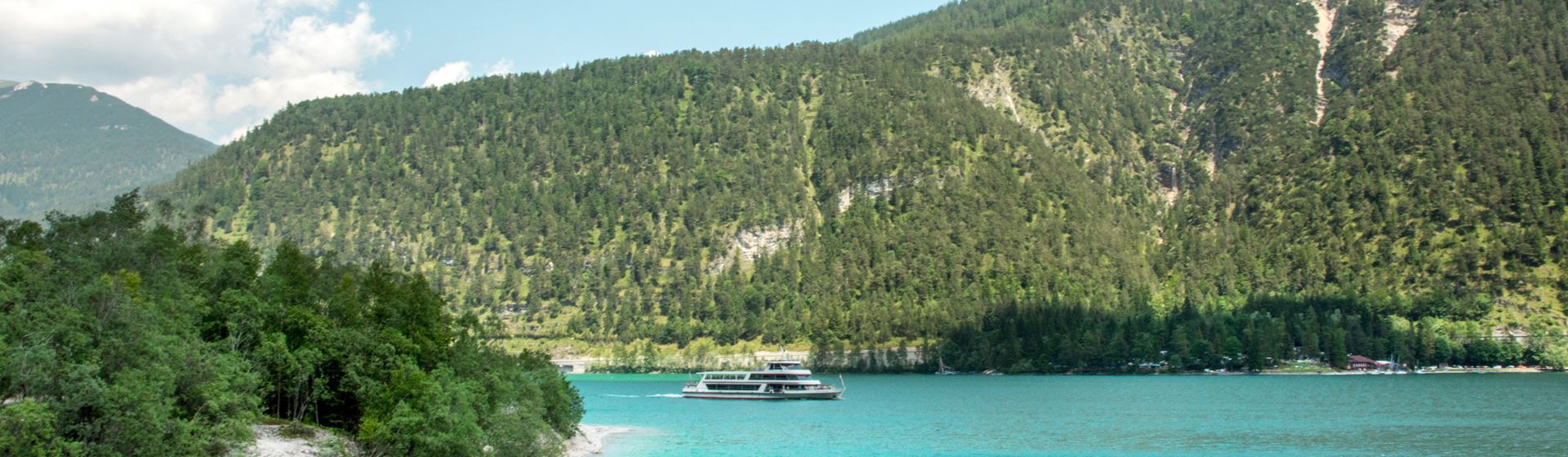 Een boot vaart op de blauwe Achensee in Oostenrijk. Op de achtergrond zijn een begroeide heuvel en een blauwe lucht te zien.