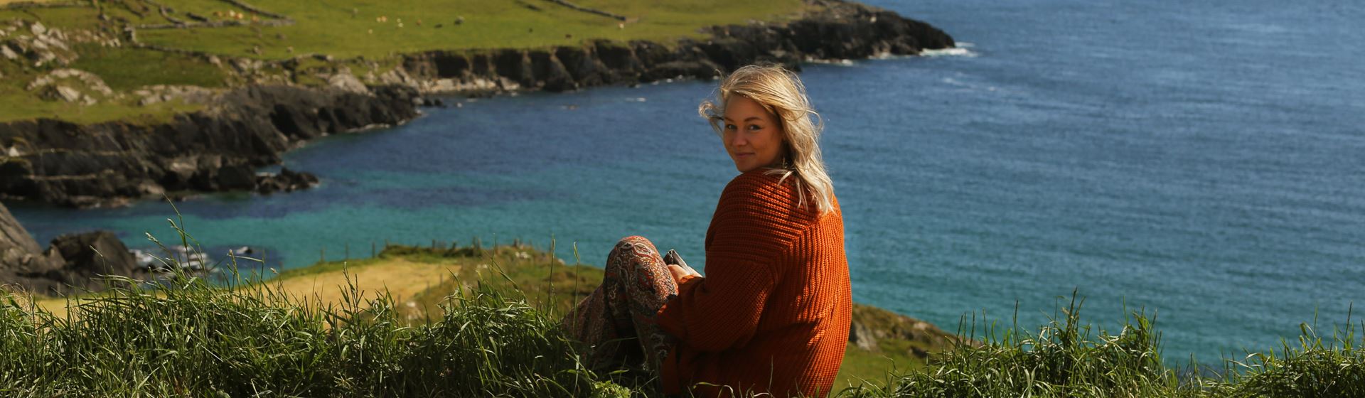Een vrouw kijkt in de lens. Ze zit in Ierland op een groene heuvel. Op de achtergrond zijn water en heuvels te zien.